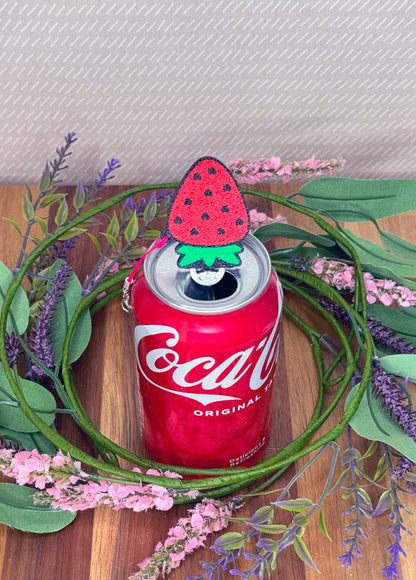 Coca-Cola can with a red strawberry Tab Buddy sitting atop a wooden surface with greenery and flowers.
