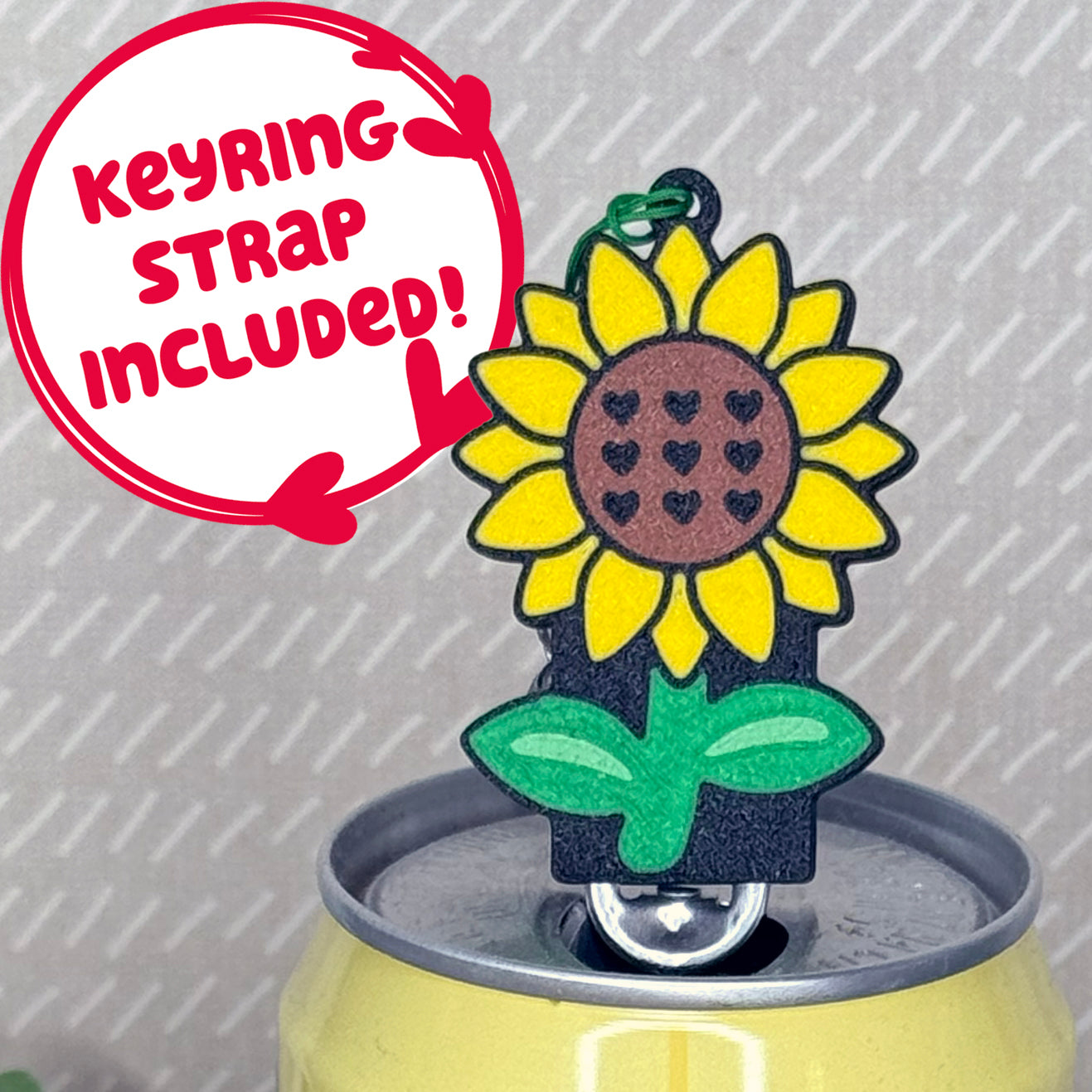 A yellow, brown and green sunflower-shaped Tab Buddy soda can opener rests atop the tab of an open can of sparkling water with a beige background. Text reads, "Keyring strap included!"