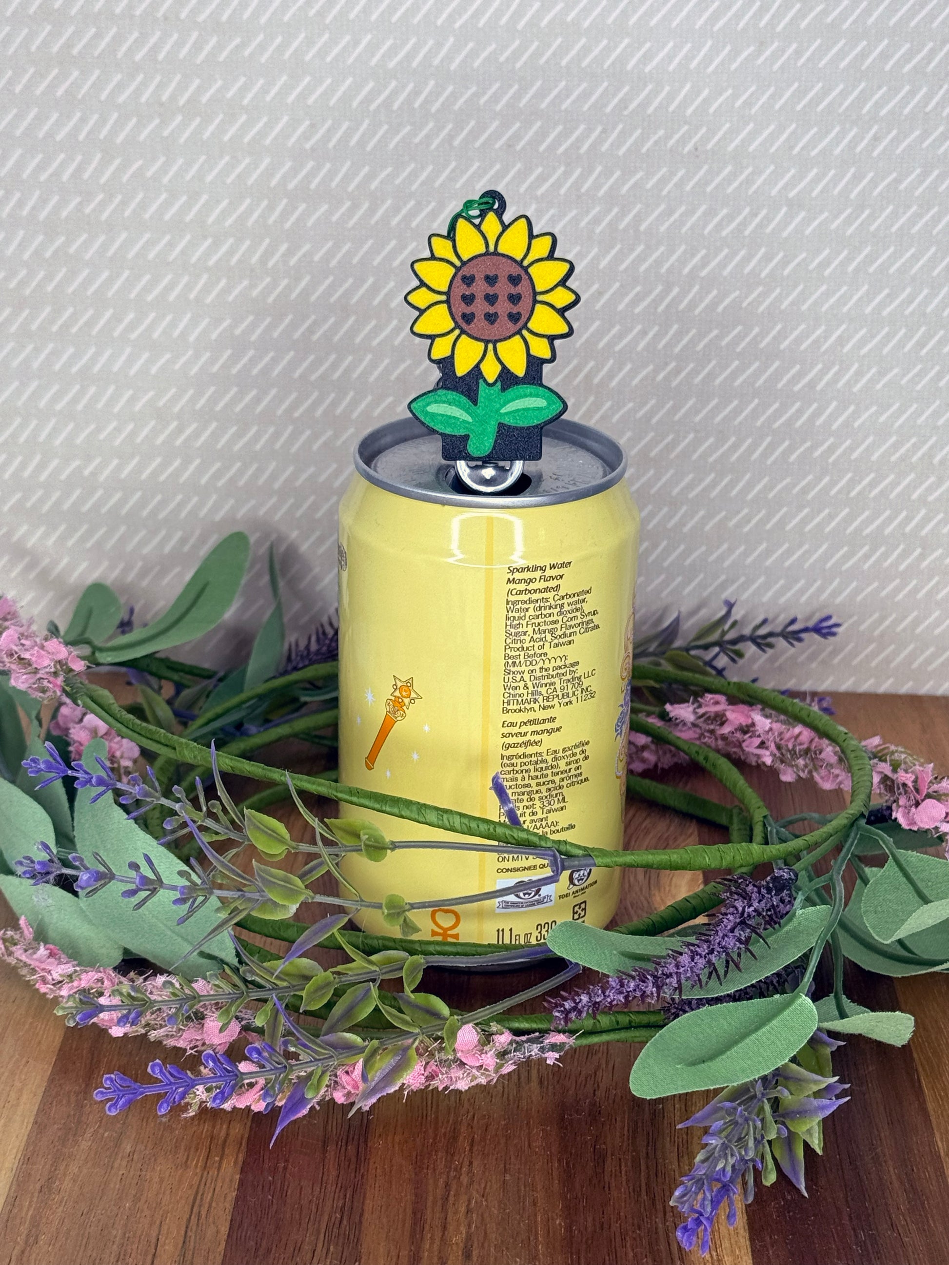A yellow, brown and green sunflower-shaped Tab Buddy soda can opener rests atop the tab of an open can of sparkling water with a beige background and floral accents.