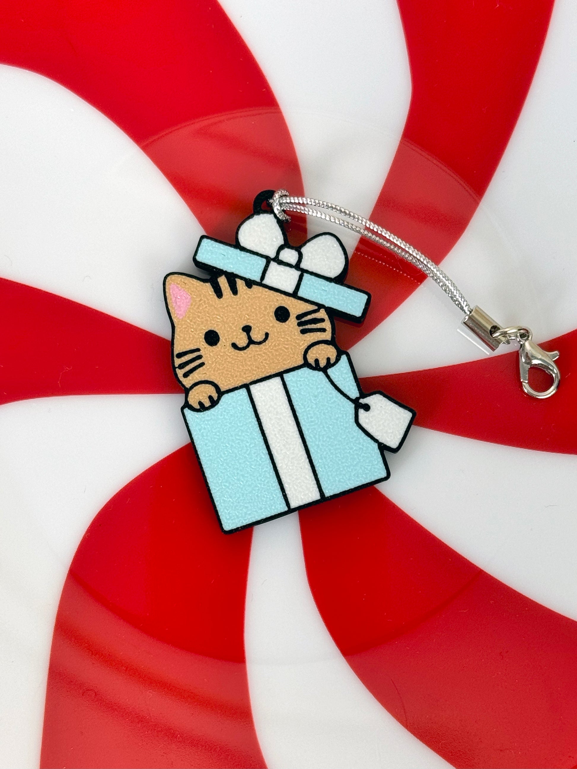 Tab Buddy keychain with a light brown cartoon cat peeking out of a blue and white gift box on a red and white striped background