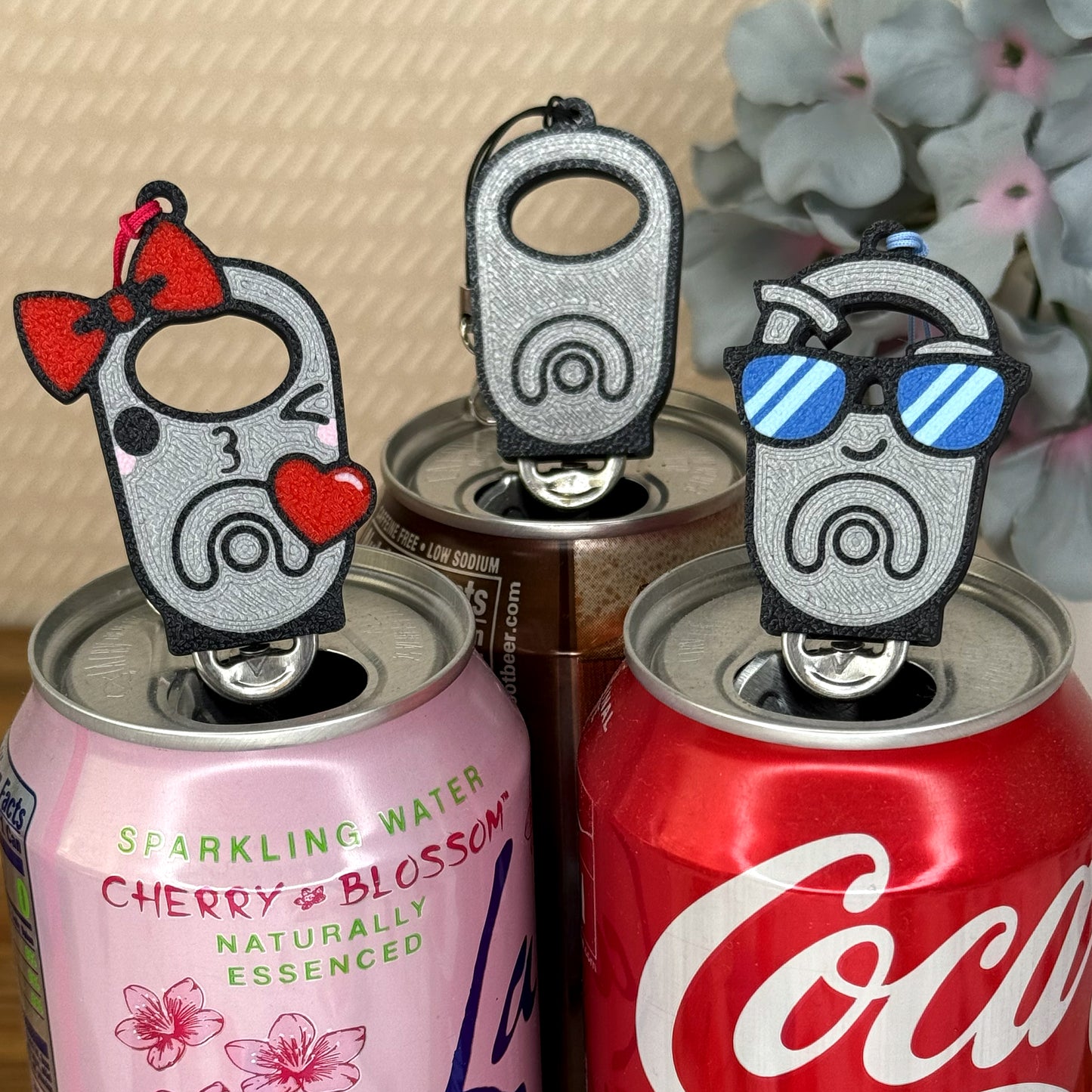 Three silver Tab Buddies rest atop soda cans with a blurred floral background.