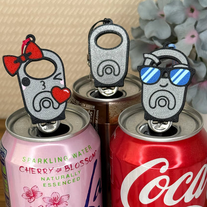 Three silver Tab Buddies rest atop soda cans with a blurred floral background.