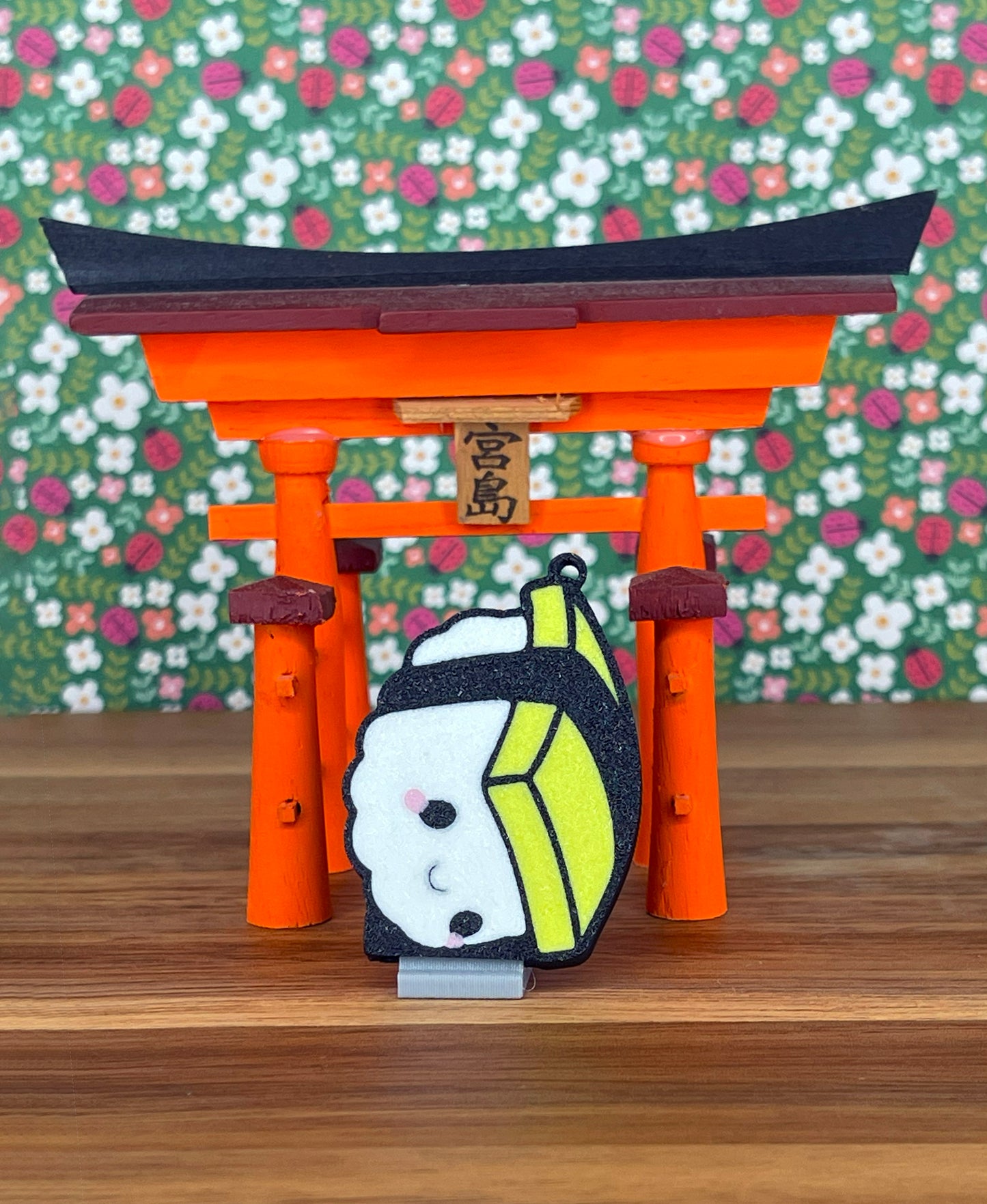 A yellow, white and black Tamago sushi Tab Buddy sits below a Japanese Toori-gate style decoration with a colorful green background of flowers and ladybugs.