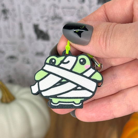 Hand holding a Halloween-themed mummy frog  Tab Buddy keychain with pumpkins in the background