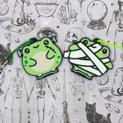 Two Halloween-themed green frog Tab Buddy keychains on a patterned background with skeletons and crystals.