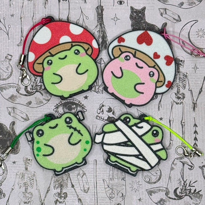 Set of four cartoon-style Tab Buddy frog keychains with different designs on a patterned background.