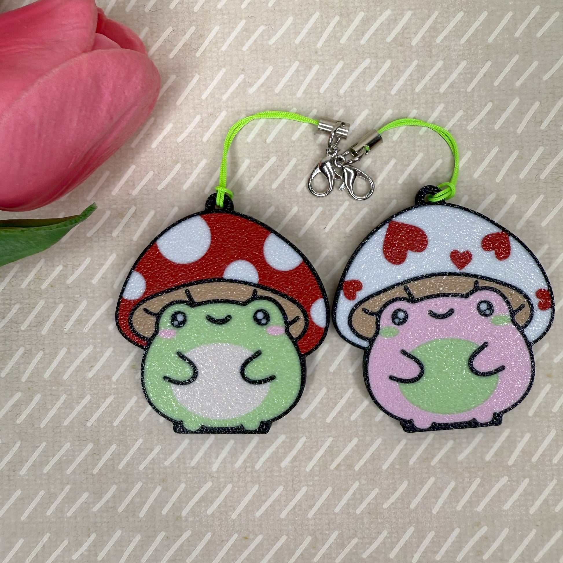 Two cartoon mushroom-shaped frogs Tab Buddies on a textured surface with a pink flower.