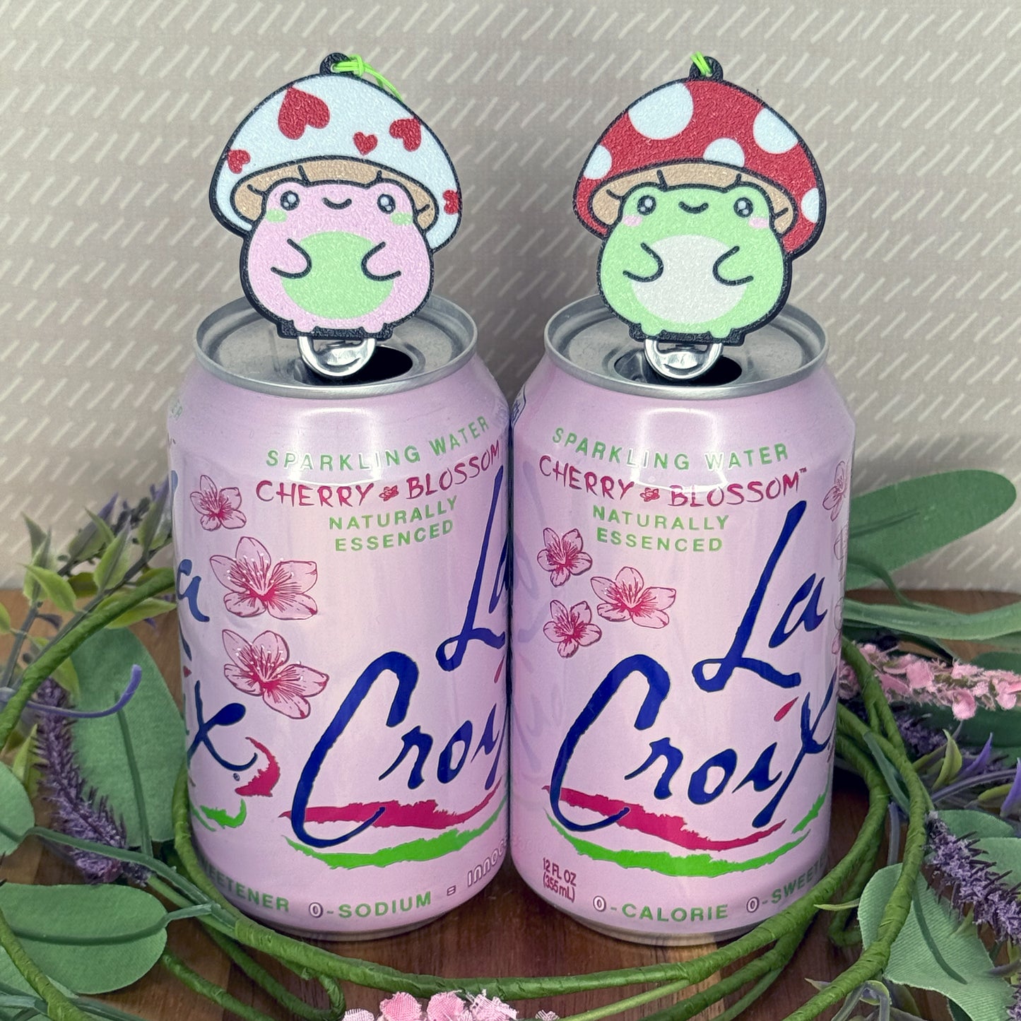 Two La Croix cherry blossom sparkling water cans with Tab Buddies of toadstool frogs on a decorative background.