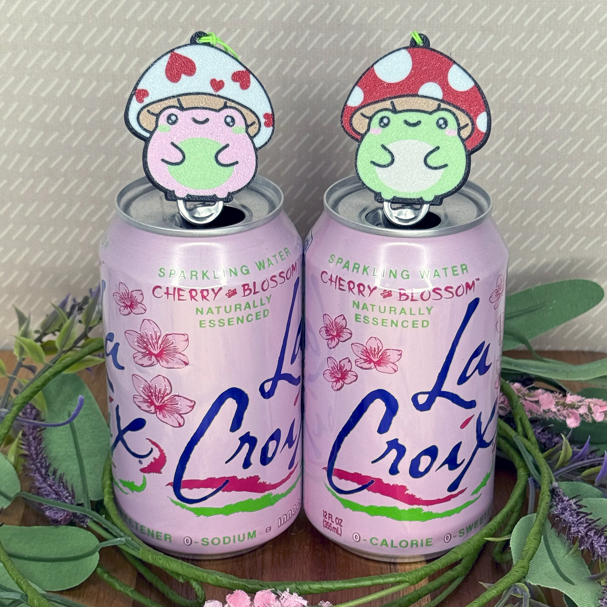 Two La Croix cherry blossom sparkling water cans with Tab Buddies of toadstool frogs on a decorative background.