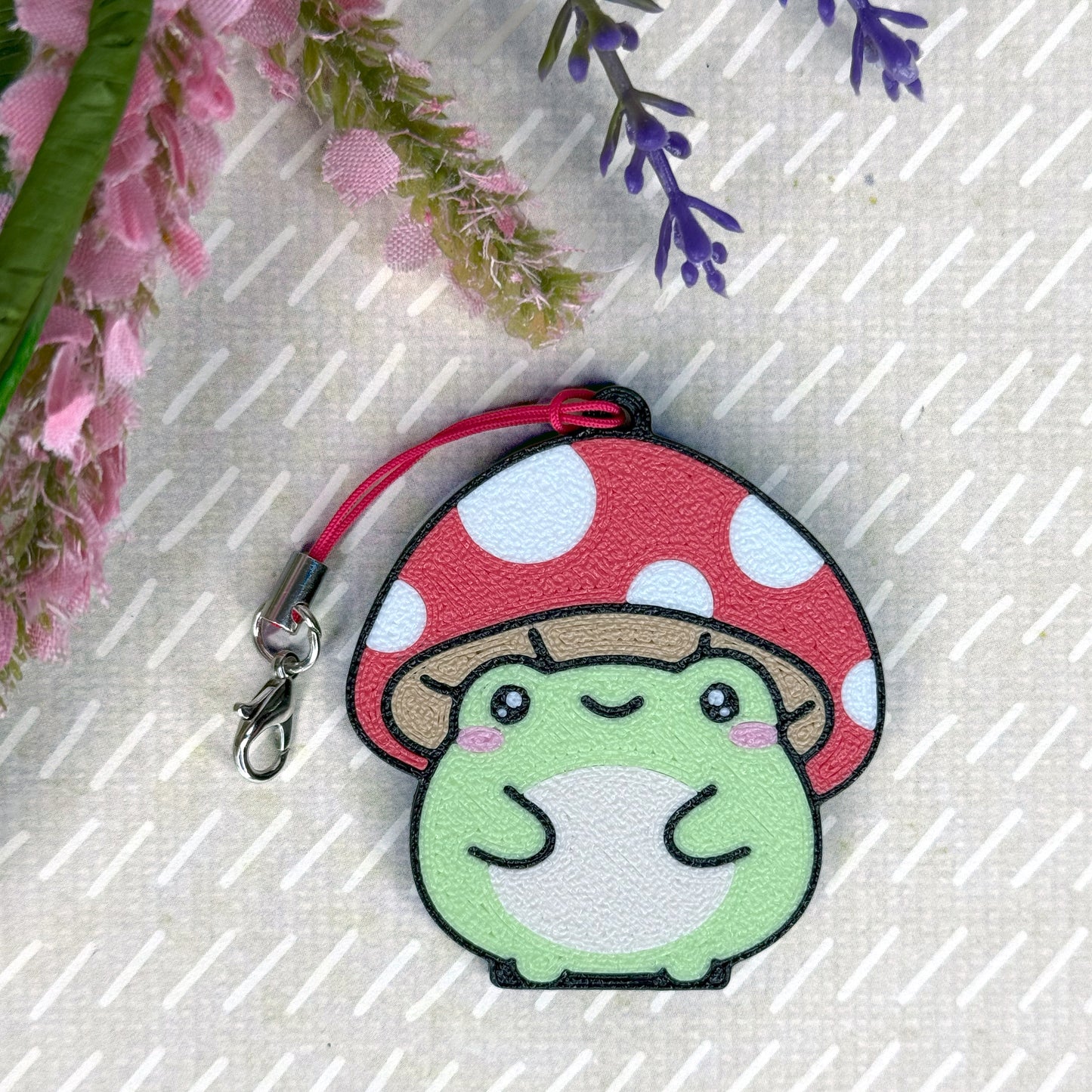 Cute green frog Tab Buddy with red polka dot mushroom on a textured surface with flowers.