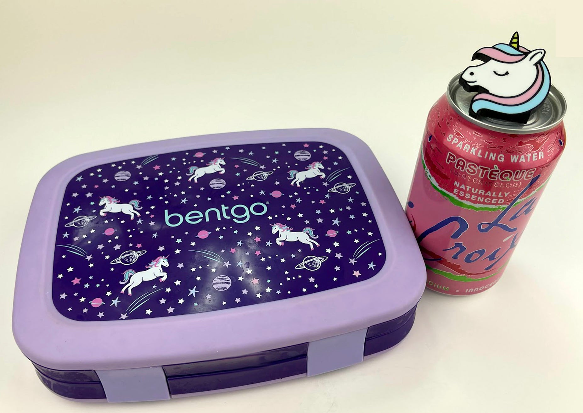 Purple lunch box with unicorn design Tab Buddy next to a pink and blue can of sparkling water.