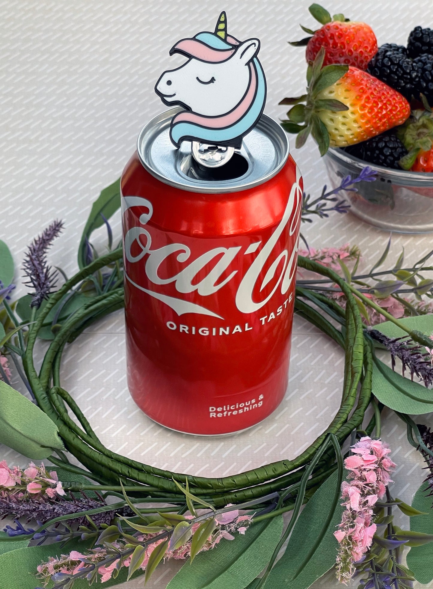 Soda can with a unicorn Tab Buddy on a decorative surface with flowers and fruits.