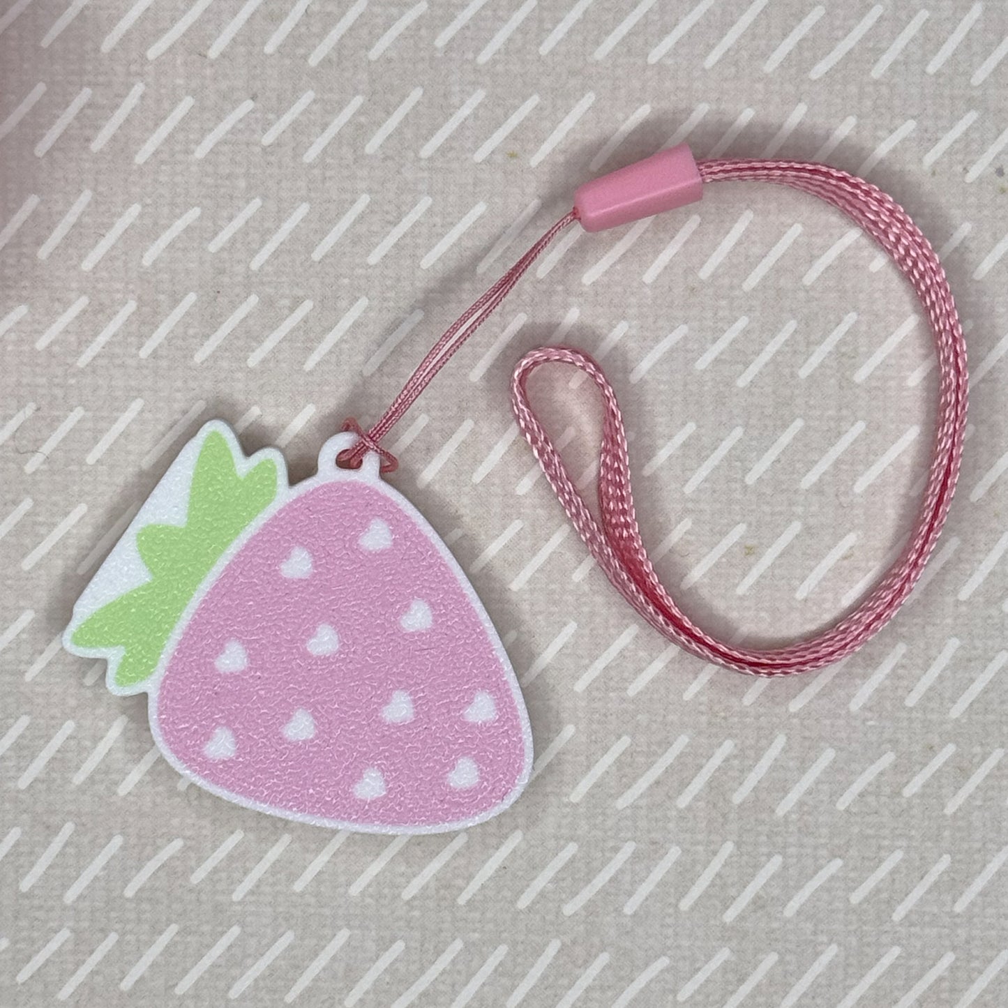 Pink strawberry-shaped Tab Buddy is shown attached to a matching pink wrist strap on a textured beige background