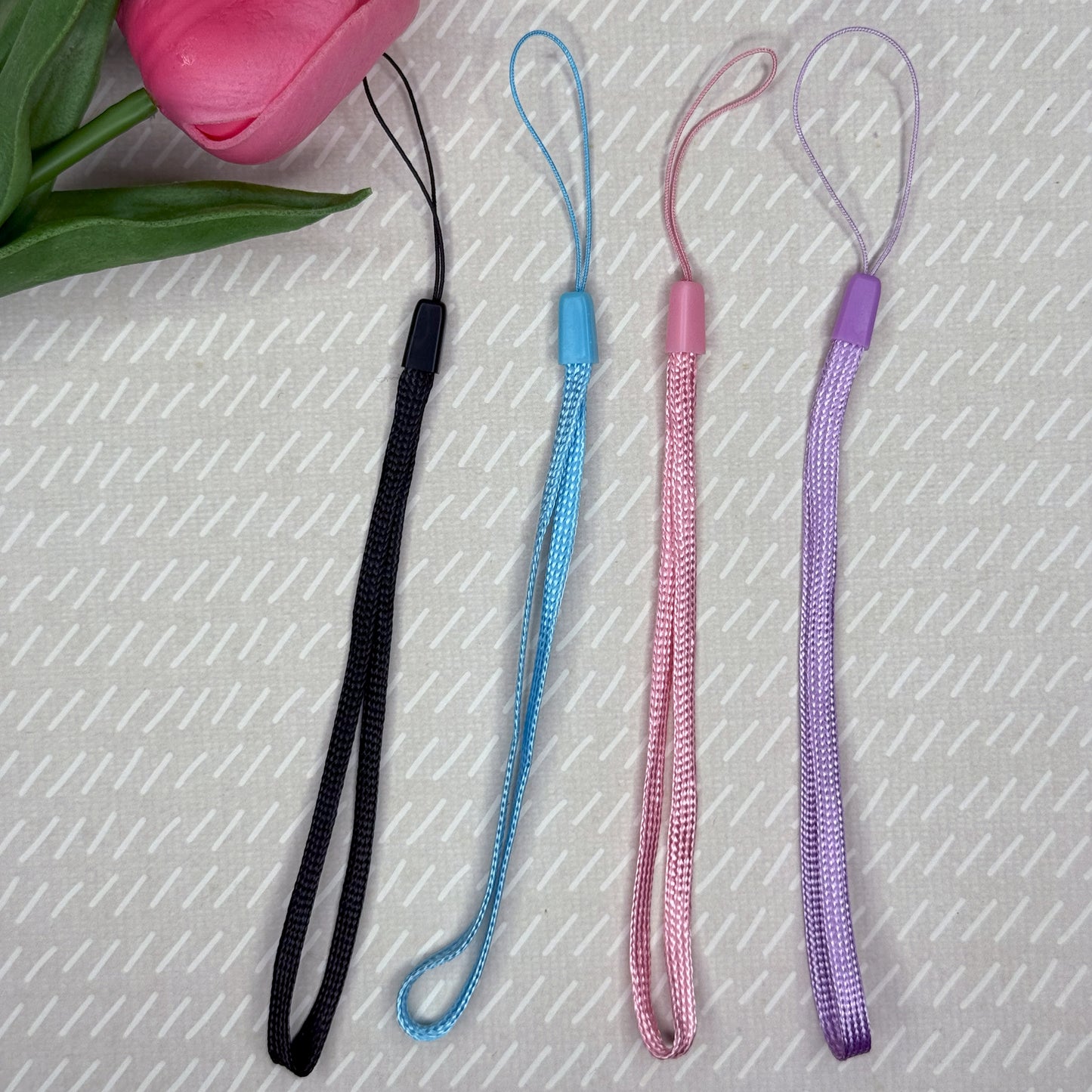 Four colorful lanyards in black, blue, pink and purple rest on a textured surface with a pink tulip.
