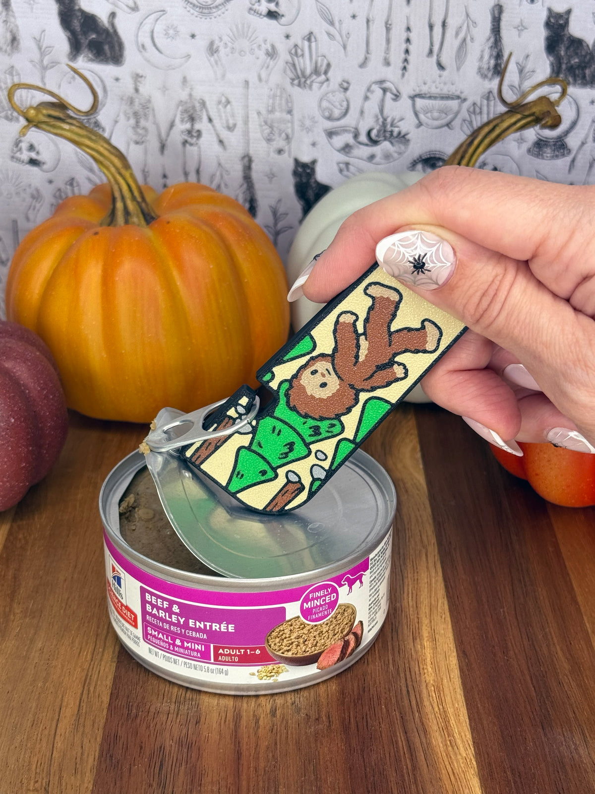 A brown and tan Bigfoot-themed Tab Buddy XL is used to peel back the lid on a can of dog food resting on a wooden table. A gothic grey backdrop and small pumpkins decorate the background.