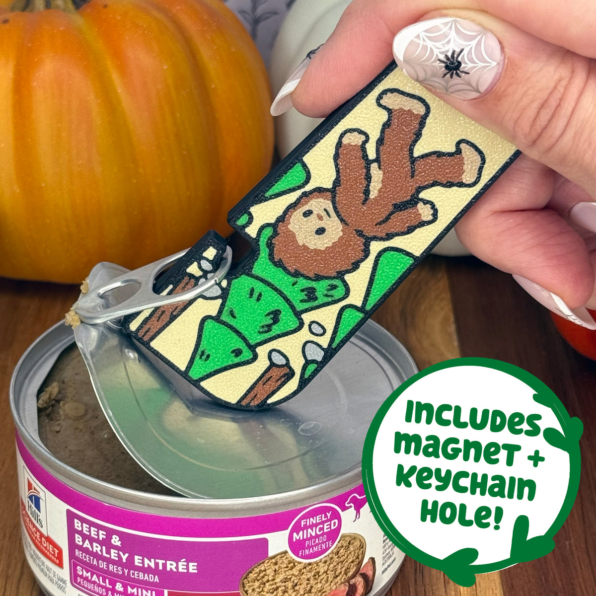 A brown and tan bigfoot-themed Tab Buddy XL is used to peel back the lid on a can of dog food. Text bubble reads, "Includes magnet + keychain hole!"