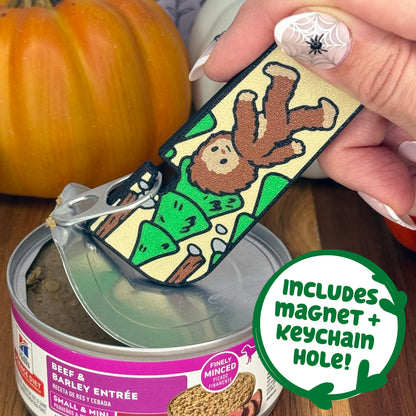 A brown and tan bigfoot-themed Tab Buddy XL is used to peel back the lid on a can of dog food. Text bubble reads, "Includes magnet + keychain hole!"