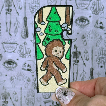 A brown and green Bigfoot-themed Tab Buddy XL is held up against a grey gothic backdrop. The XL design features a brown sasquatch with tan background and green trees.
