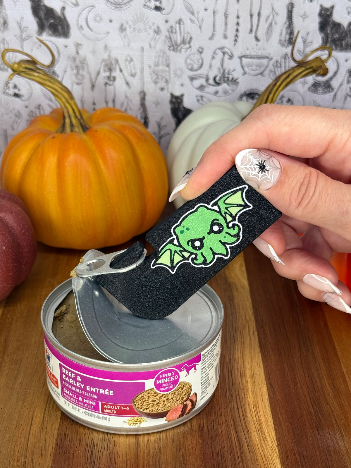 A black and green Cthulhu-themed Tab Buddy XL is used to peel back the lid on a can of dog food resting on a wooden table. A gothic grey backdrop and small pumpkins decorate the background.