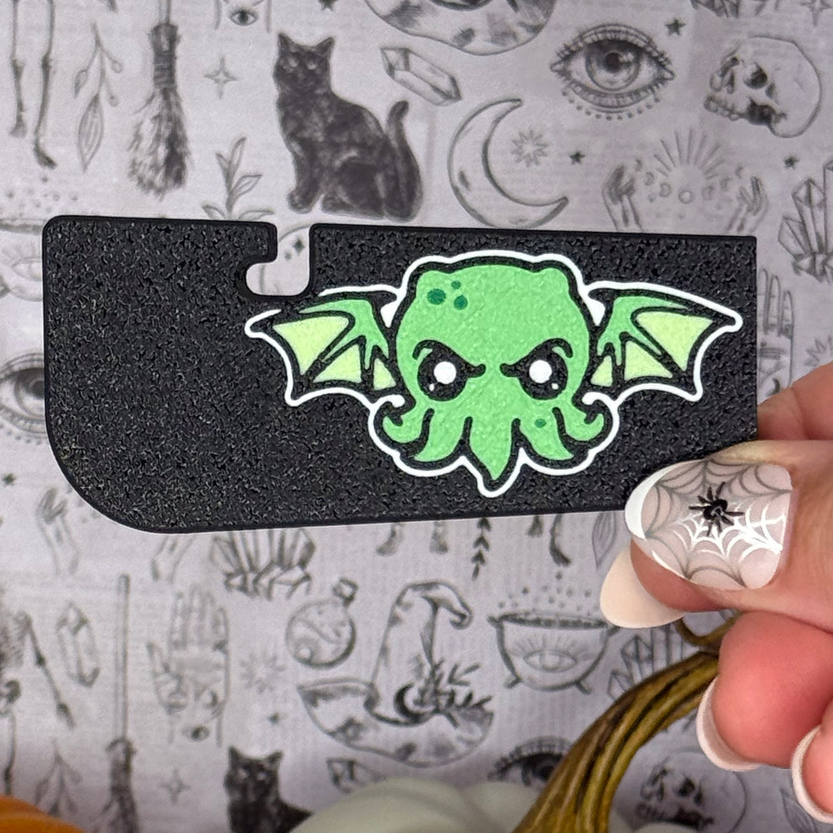 A black cthulhu-themed Tab Buddy XL is held up against a grey and black gothic backdrop. The cthulhu is green with short tentatles and pointed wings.