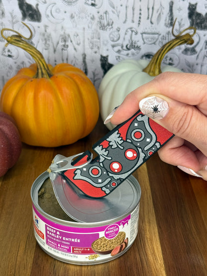 A grey and red Mothman-themed Tab Buddy XL is used to peel back the lid on a can of dog food resting on a wooden table. A gothic grey backdrop and small pumpkins decorate the background.