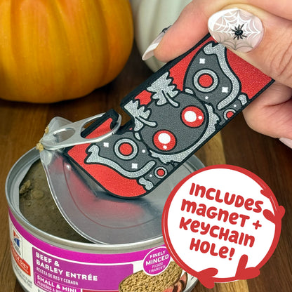 A grey and red Mothman-themed Tab Buddy XL is used to peel back the lid on a can of dog food. Text bubble reads, "Includes magnet + keychain hole!"