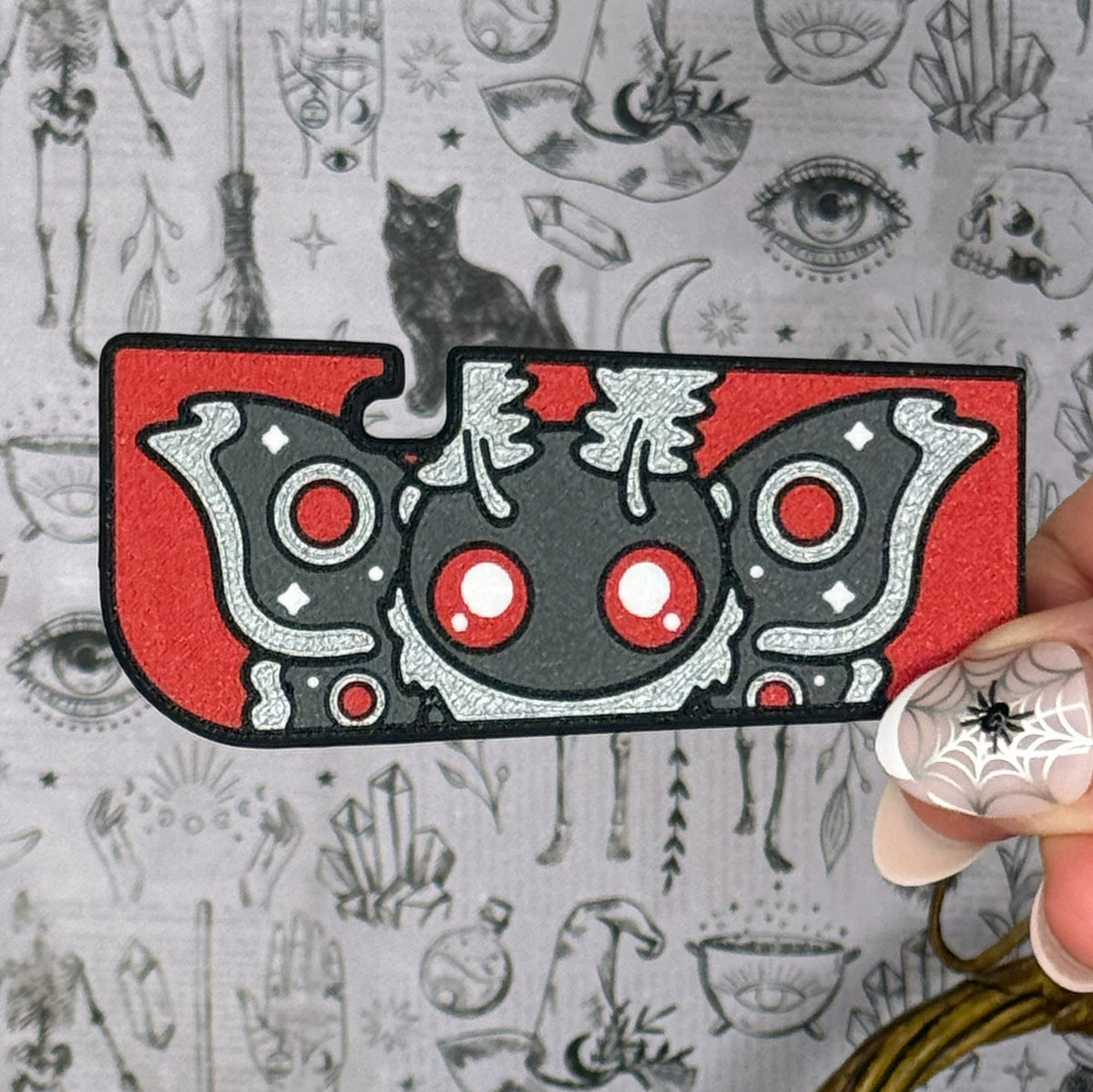 A red and grey mothman-themed Tab Buddy XL is held up against a grey gothic backdrop. The XL design features a grey mothman with dark grey wings, red eyes and silver accents.