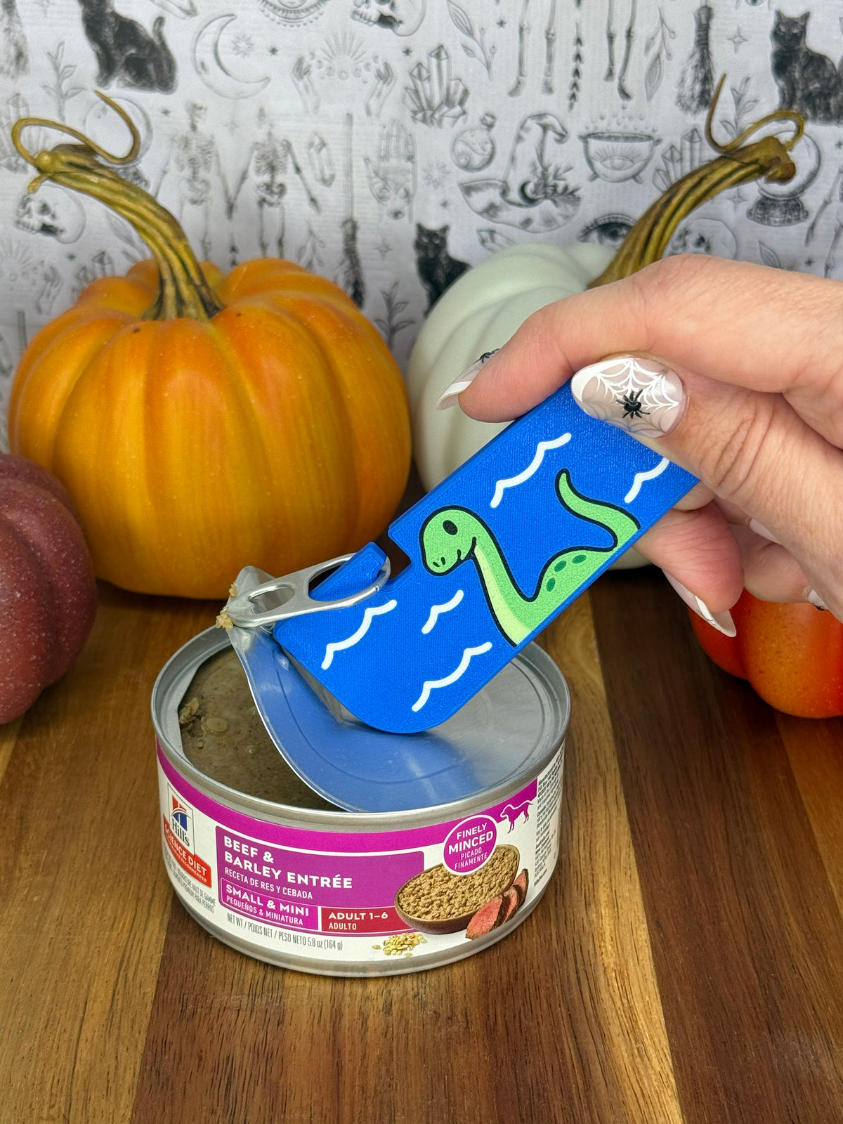 A blue loch ness monster-themed Tab Buddy XL is used to peel back the lid on a can of dog food resting on a wooden table. A gothic grey backdrop and small pumpkins decorate the background.