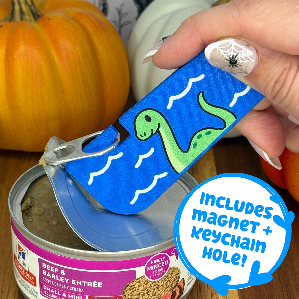 A blue loch ness monster-themed Tab Buddy XL is used to peel back the lid on a can of dog food. Text bubble reads, "Includes magnet + keychain hole!"