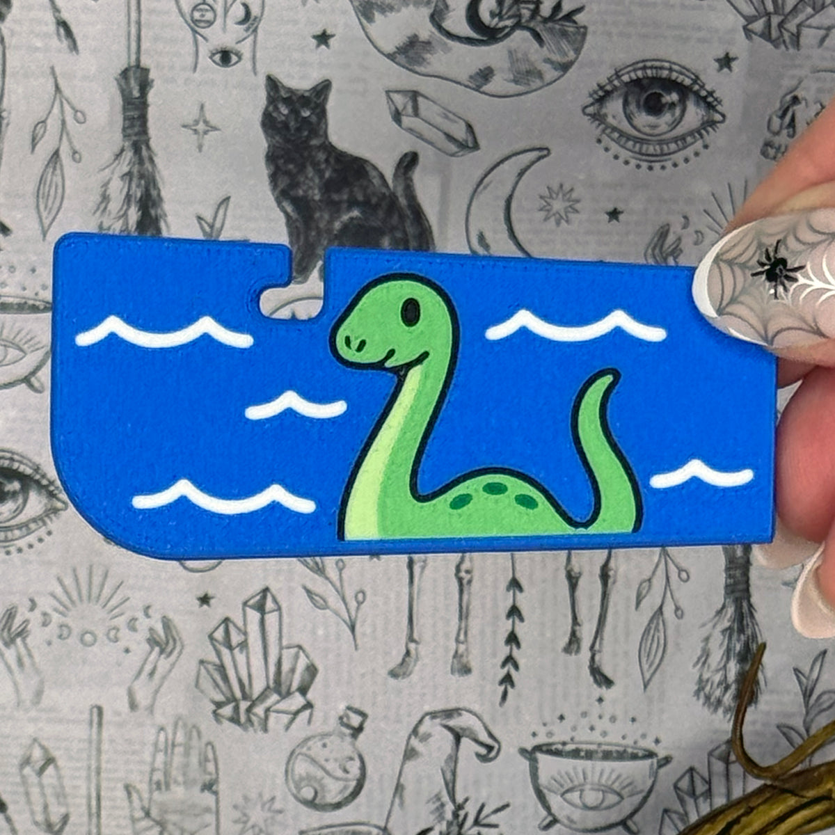 A blue Loch Ness Monster-themed Tab Buddy XL is held up against a grey gothic backdrop. The XL design features a green Nessie with dark green spots swimming in a blue lake with gentle waves. 