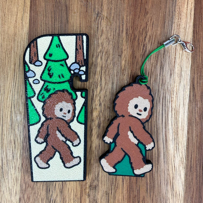 A brown and tan Bigfoot-themed Tab Buddy XL and its matching soda-sized Tab Buddy with keychain strap rest on a wooden surface.