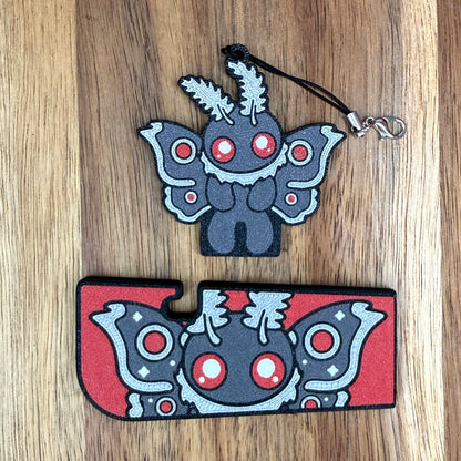 A grey and red Mothmani-themed Tab Buddy XL and its matching soda-sized Tab Buddy with keychain strap rest on a wooden surface.