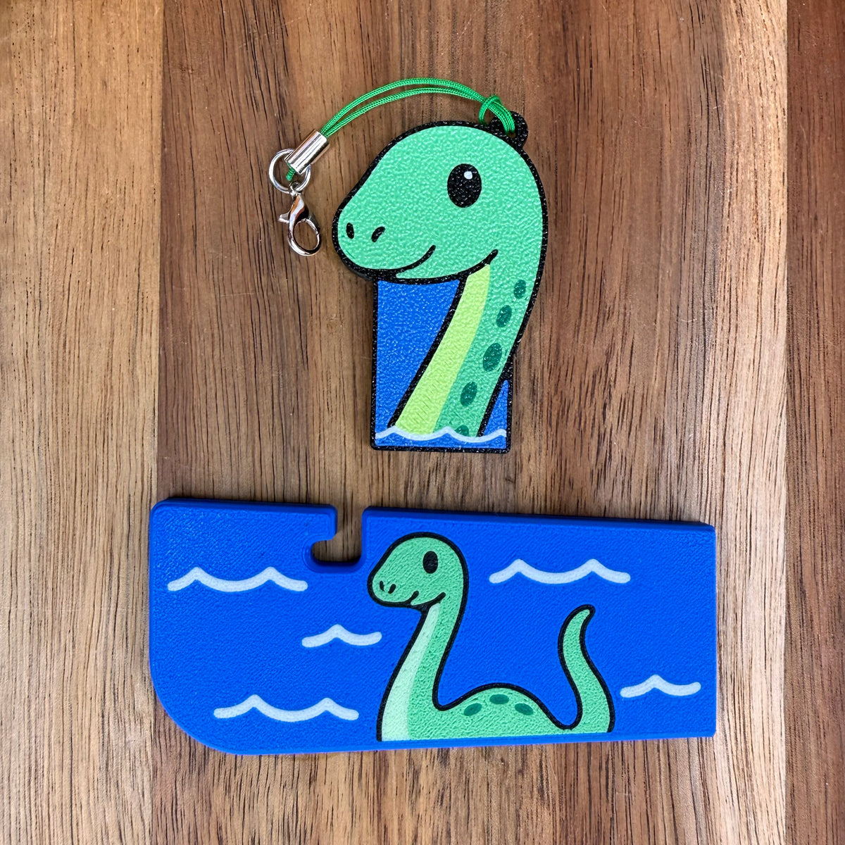 A green and  blue Loch Ness Monster-themed Tab Buddy XL and its matching soda-sized Tab Buddy with keychain strap rest on a wooden surface.