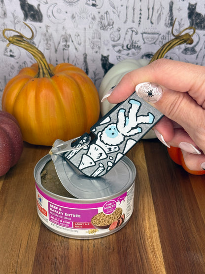 A silver and white Yeti-themed Tab Buddy XL is used to peel back the lid on a can of dog food resting on a wooden table. A gothic grey backdrop and small pumpkins decorate the background.