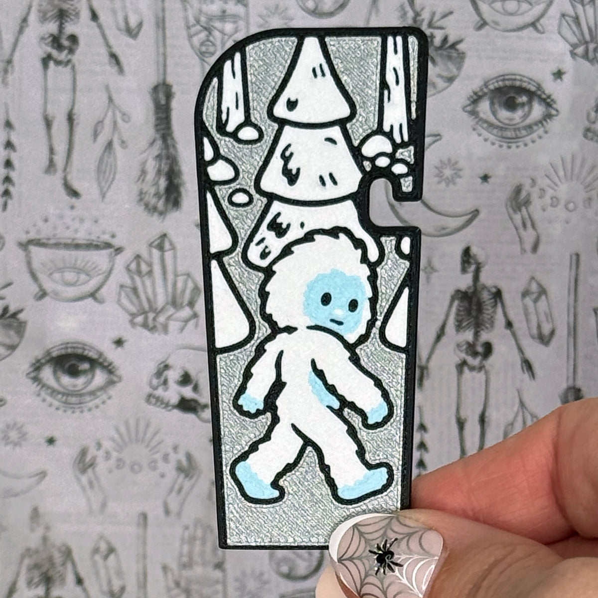 A silver and white snow yeti-themed Tab Buddy XL is held up against a grey gothic backdrop. The XL design features a white and blue abominable snowman with a silvery background and snowy white trees.