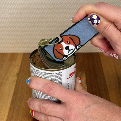 Hand using a Tab Buddy XL with a Beagle dog design to open a can of dog food on a wooden surface.