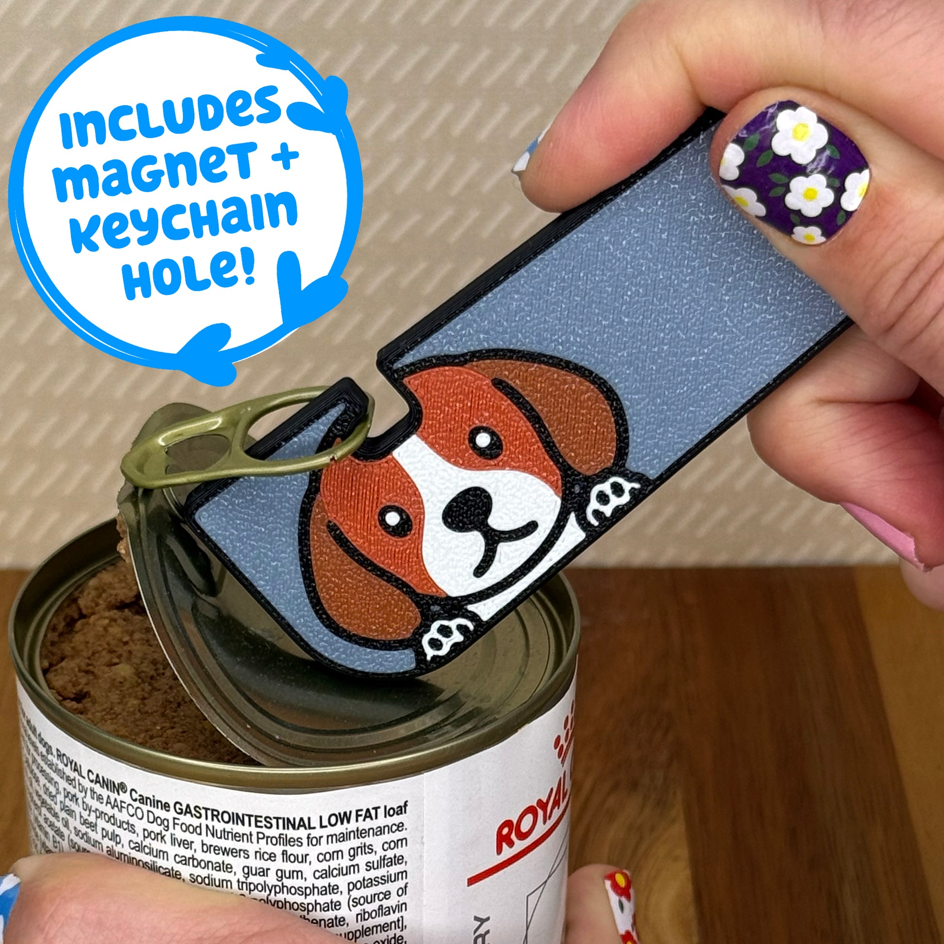 Hand using a Tab Buddy XL with a Beagle dog design to open a can of dog food on a wooden surface. Text bubble reads, "Includes magnet + keychain strap!"