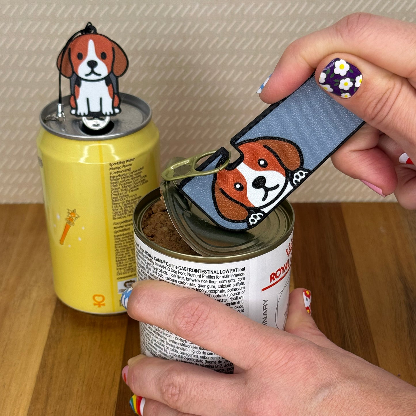 A person opens a can of dog food with a Beagle-designed Tab Buddy XL. In the background, a matching soda-sized Tab Buddy rests atop the tab of a can of sparkling water.