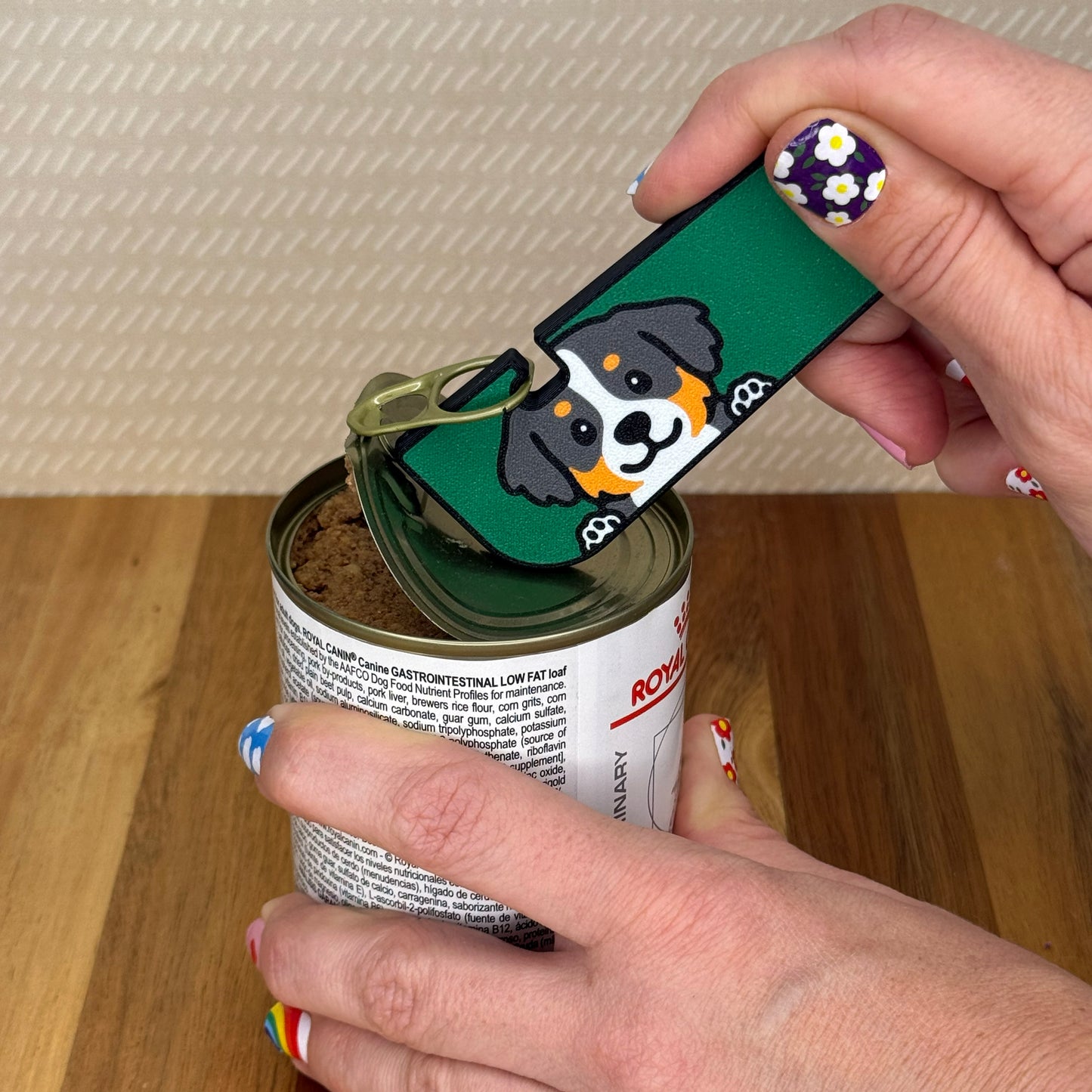Hand using a Tab Buddy XL with a Bernese Mountain dog design to open a can of dog food on a wooden surface.