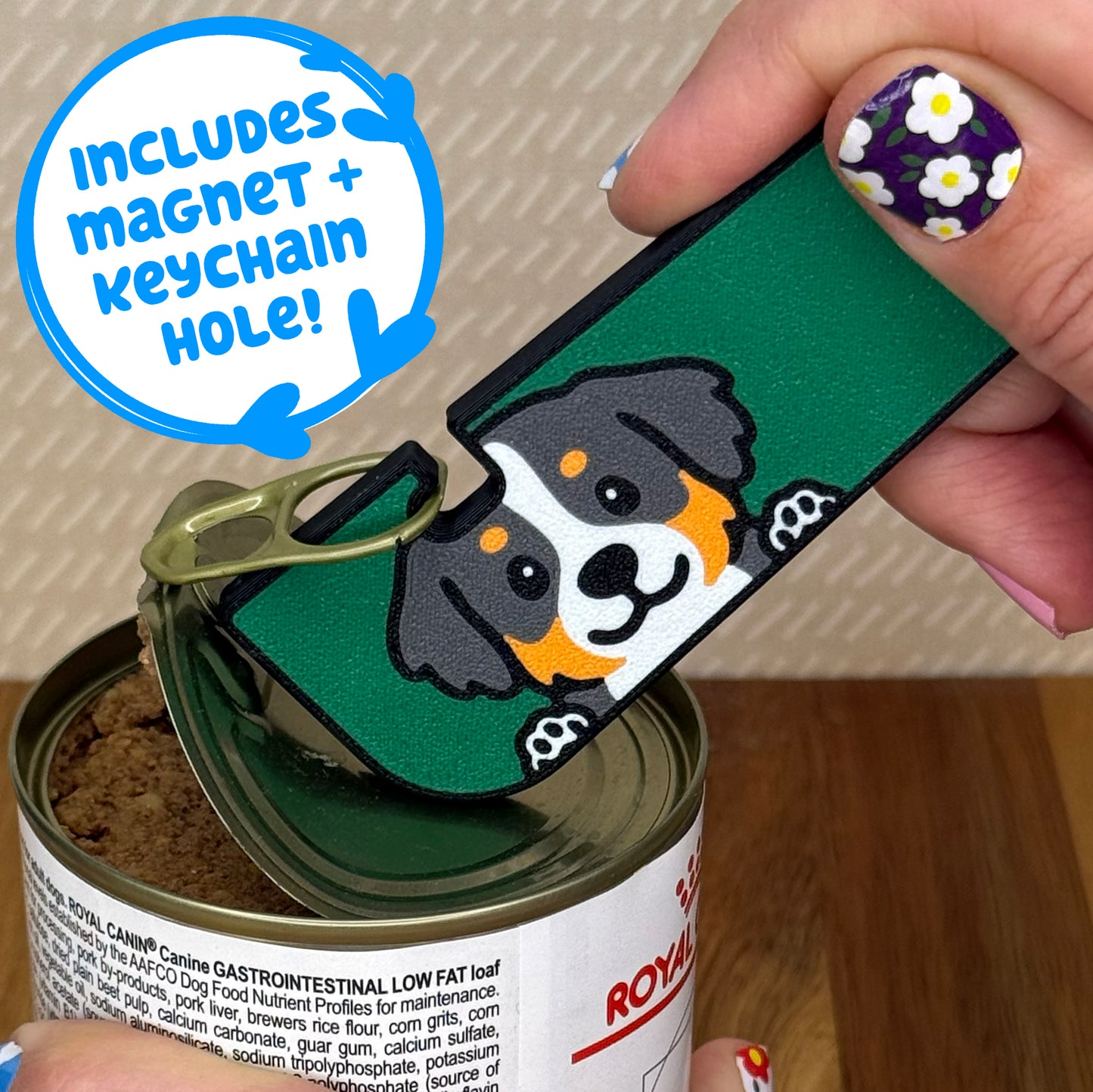 Hand using a Tab Buddy XL with a Bermese Mountain dog design to open a can of dog food on a wooden surface. Text bubble reads, "Includes magnet + keychain strap!"