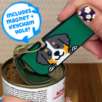Hand using a Tab Buddy XL with a Bermese Mountain dog design to open a can of dog food on a wooden surface. Text bubble reads, "Includes magnet + keychain strap!"