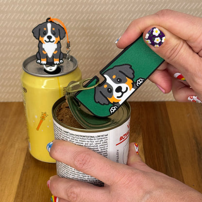 A person opens a can of dog food with a Bernese Mountain Dog-design Tab Buddy XL. In the background, a matching soda-sized Tab Buddy rests atop the tab of a can of sparkling water.