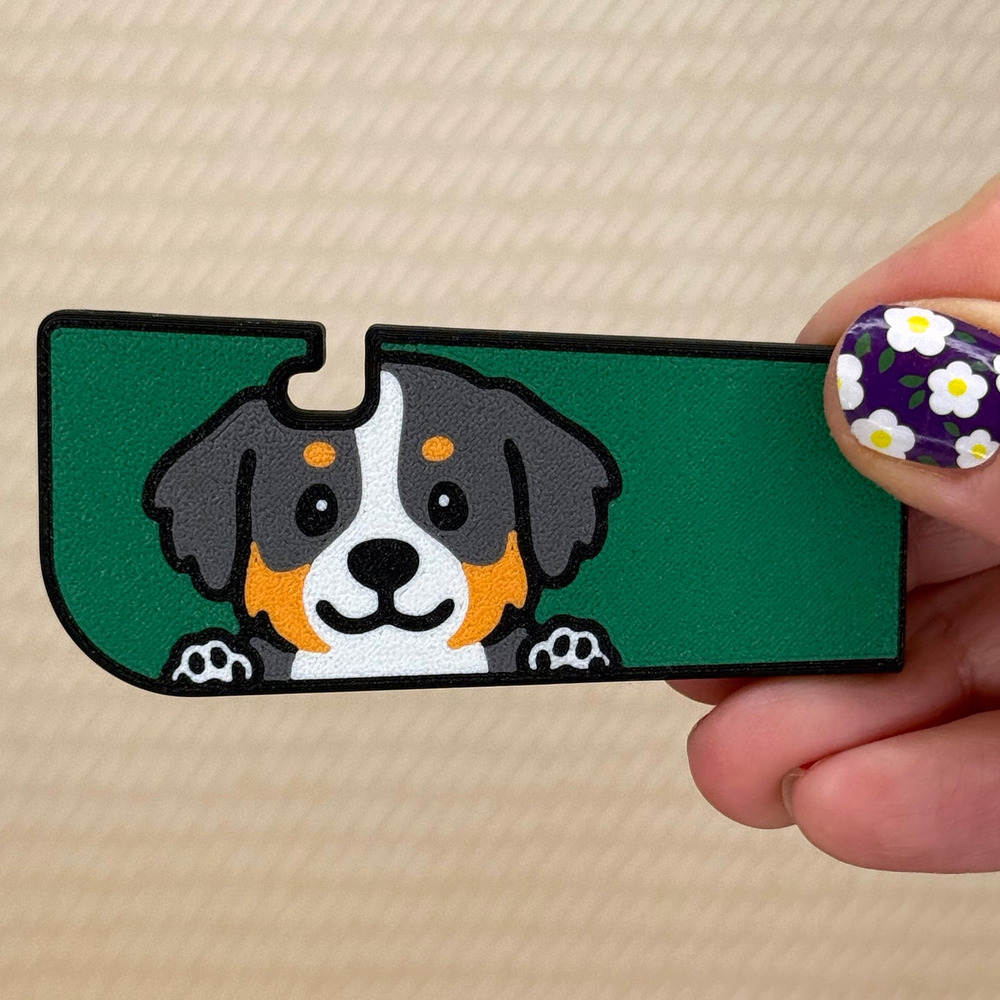 Hand holding a Tab Buddy XL with a cute Bernese Mountain Dog design and green background on its surface. A tan surface is in the background of the photo.