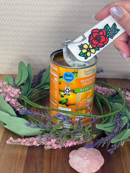 White Tab Buddy XL with Floral design of winter flowers in red, yellow and light blue is used to open a can of Sweet Harvest Mandarin Oranges. Decorative flowers surround the can against a beige background.