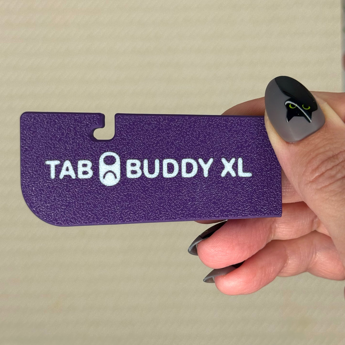 Plum purple 'TAB BUDDY XL' product held by a hand with gray nail polish against a beige background