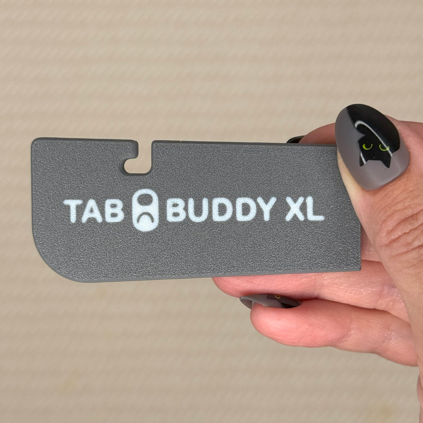 Hand holding a slate grey 'TAB BUDDY XL' product against a beige background