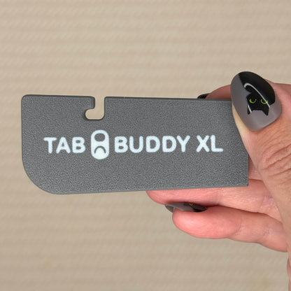 Hand holding a slate grey 'TAB BUDDY XL' product against a beige background
