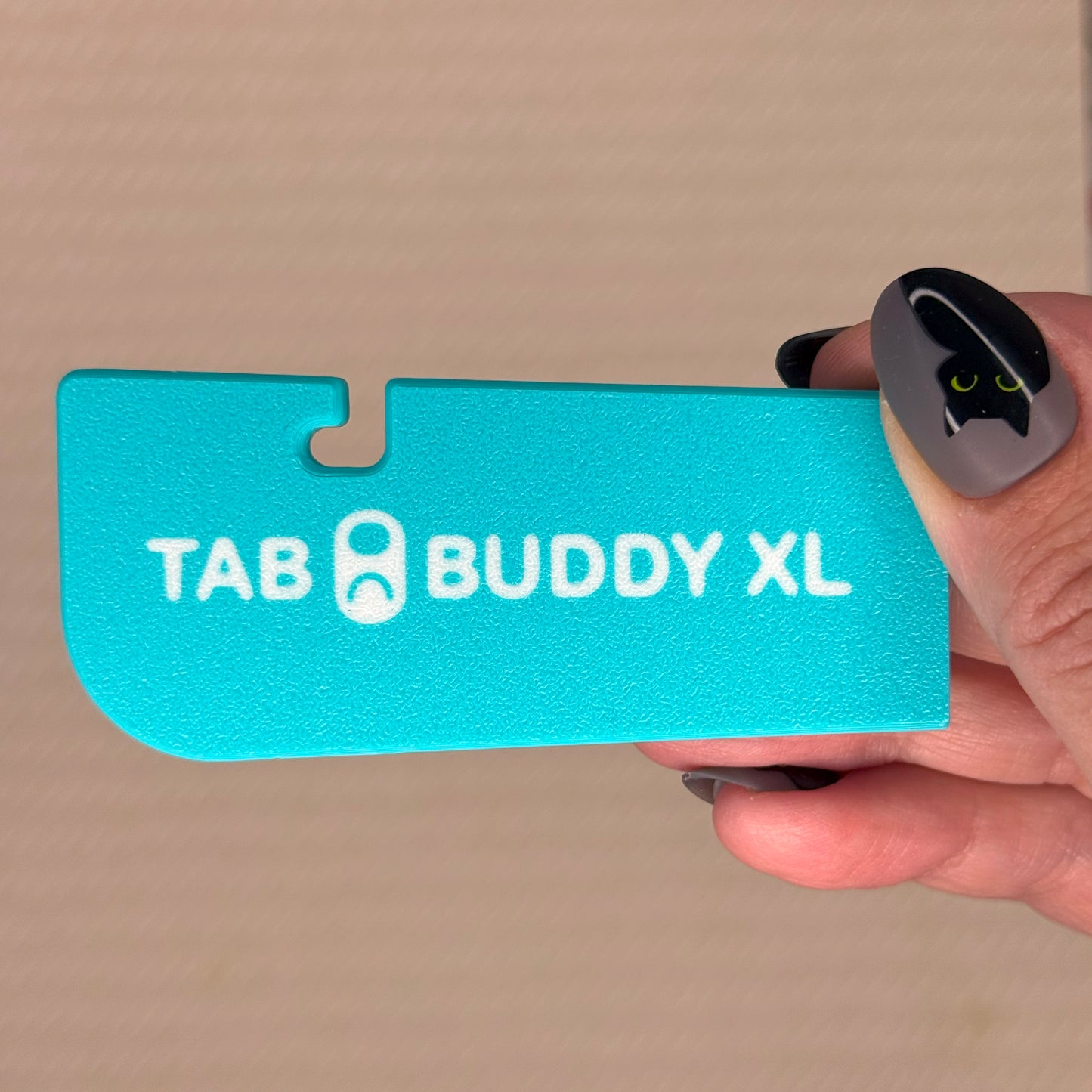 Turquoise 'TAB BUDDY XL' product held by a hand against a beige background