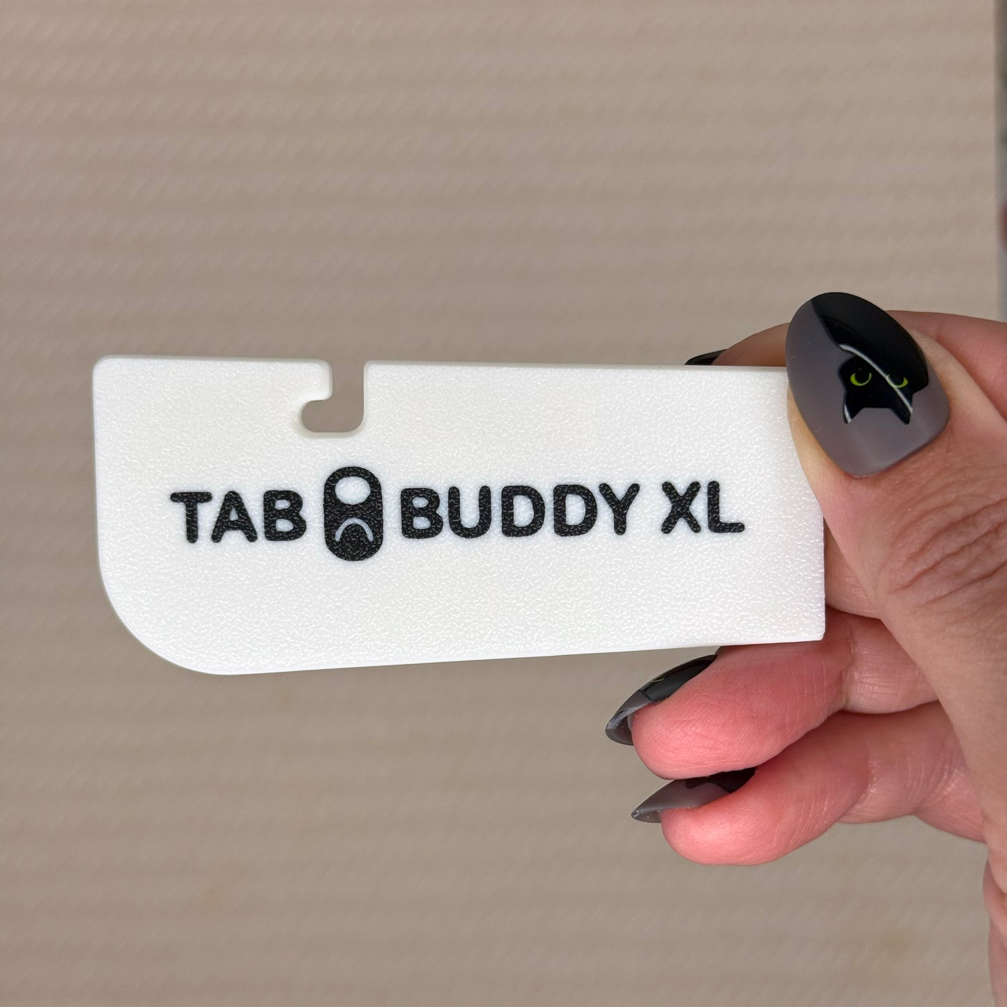 Hand holding a white 'TAB BUDDY XL' product against a beige background
