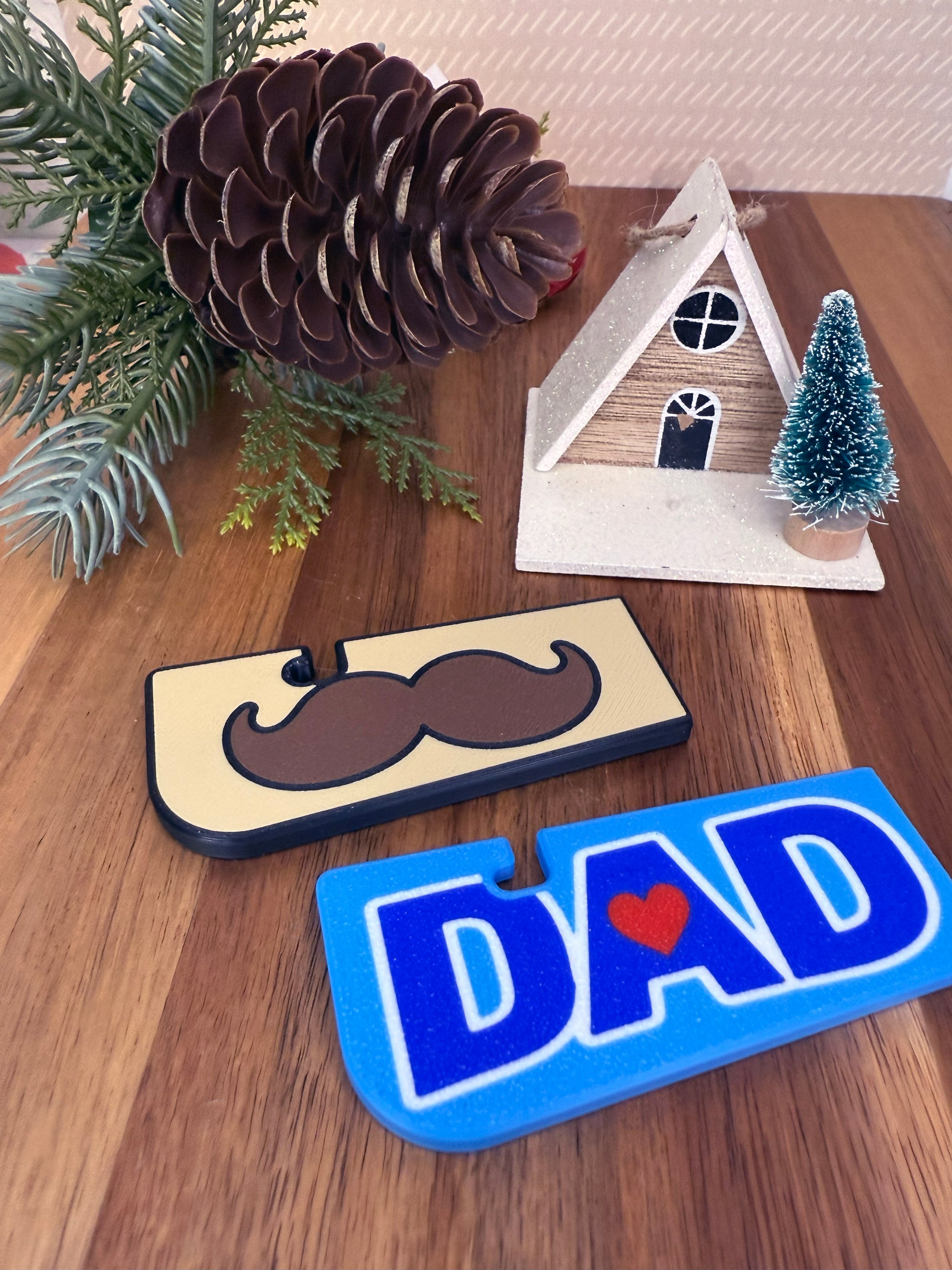 Decorative items including a mustache design and 'DAD' design Tab Buddy XL on a wooden surface with Christmas decorations.